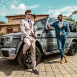 Two stylish men in tailored suits posing beside a luxury Mercedes G-Class in a sunny outdoor setting