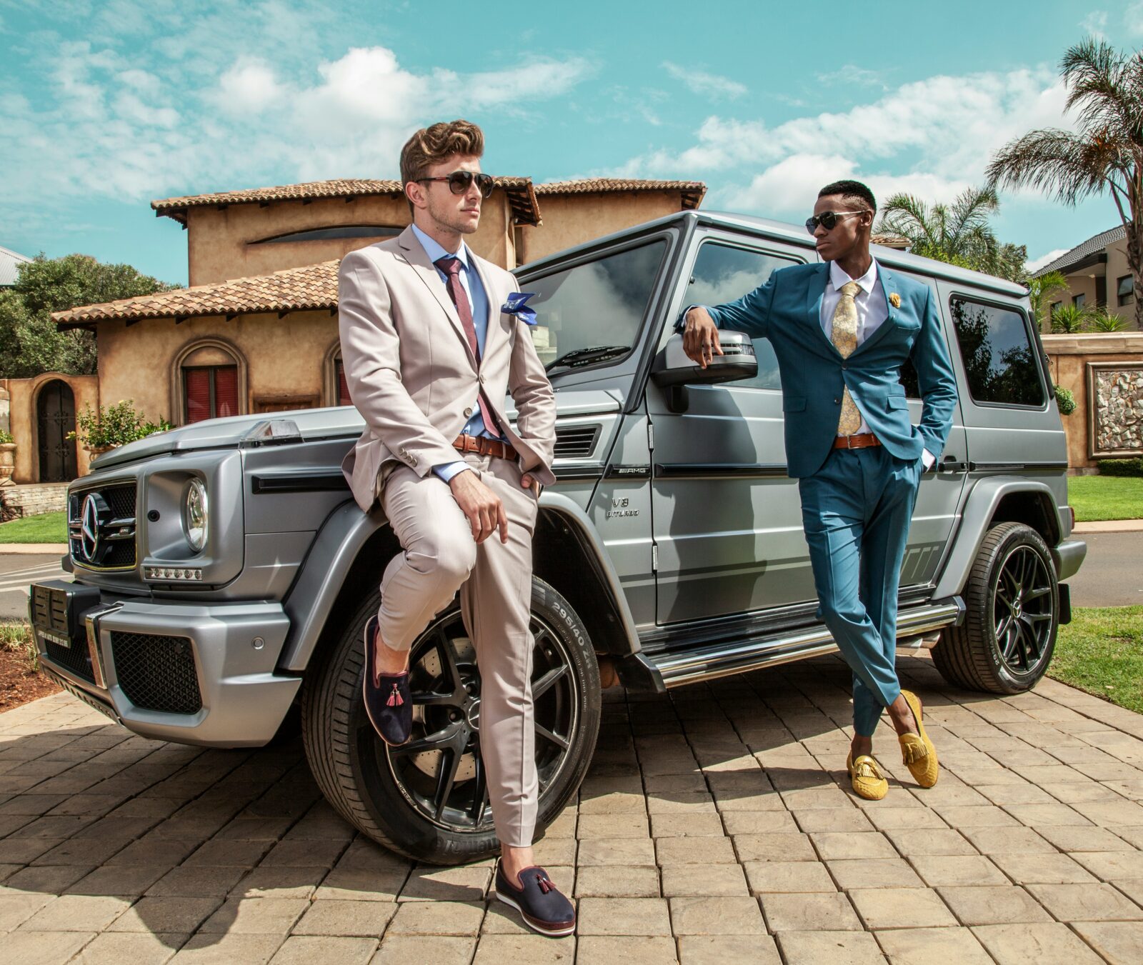 Two stylish men in tailored suits posing beside a luxury Mercedes G-Class in a sunny outdoor setting