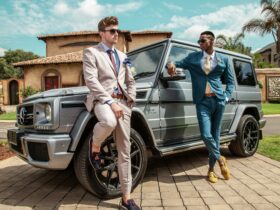 Two stylish men in tailored suits posing beside a luxury Mercedes G-Class in a sunny outdoor setting