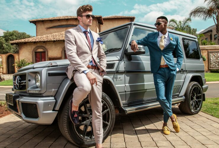Two stylish men in tailored suits posing beside a luxury Mercedes G-Class in a sunny outdoor setting